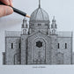 A hand holds a black pencil over a detailed black-and-white architectural drawing of Galway Cathedral on textured white paper, with a rustic wooden surface visible at the bottom.