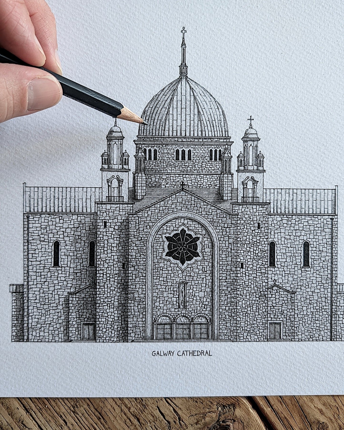 A hand holds a black pencil over a detailed black-and-white architectural drawing of Galway Cathedral on textured white paper, with a rustic wooden surface visible at the bottom.