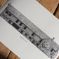 A black and white architectural art print of a stone building, Tudor Barn Eltham, shown laid flat on a rustic wooden surface with diagonal shadows.