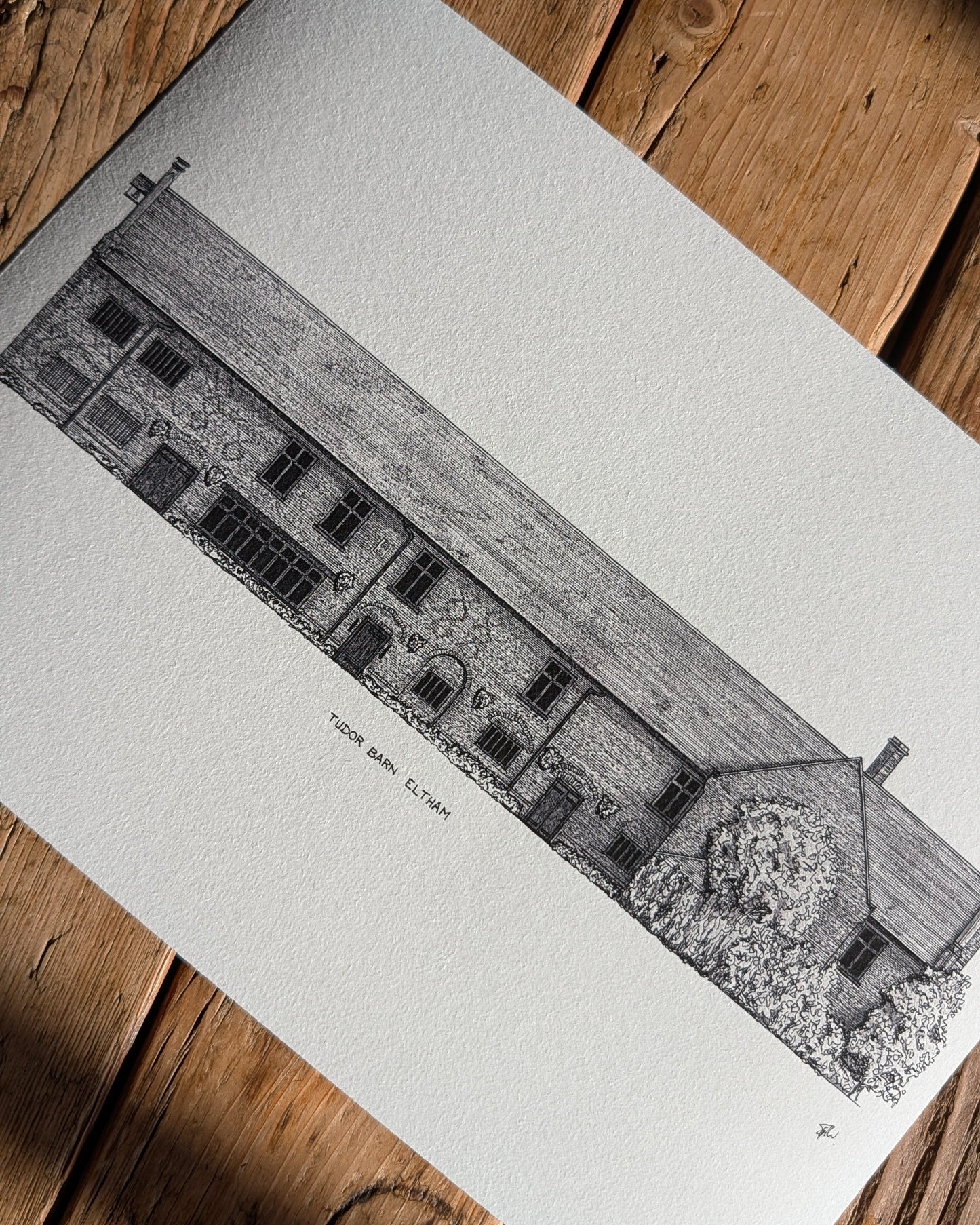 A black and white architectural art print of a stone building, Tudor Barn Eltham, shown laid flat on a rustic wooden surface with diagonal shadows.