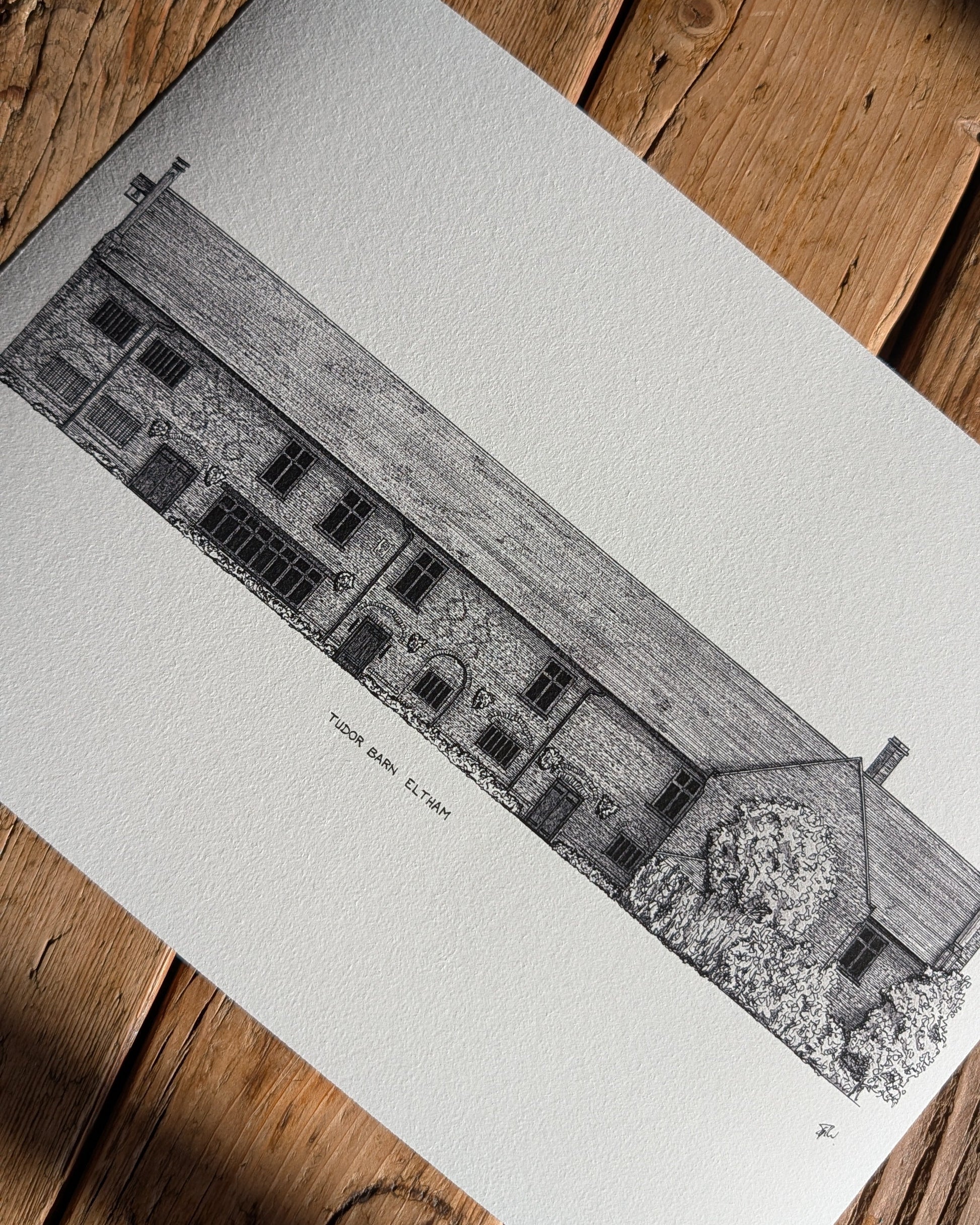 A black and white architectural art print of a stone building, Tudor Barn Eltham, shown laid flat on a rustic wooden surface with diagonal shadows.