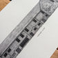 A black and white architectural sketch print of 'Tudor Barn, Eltham' on textured white paper, displayed diagonally on a rustic wooden surface.