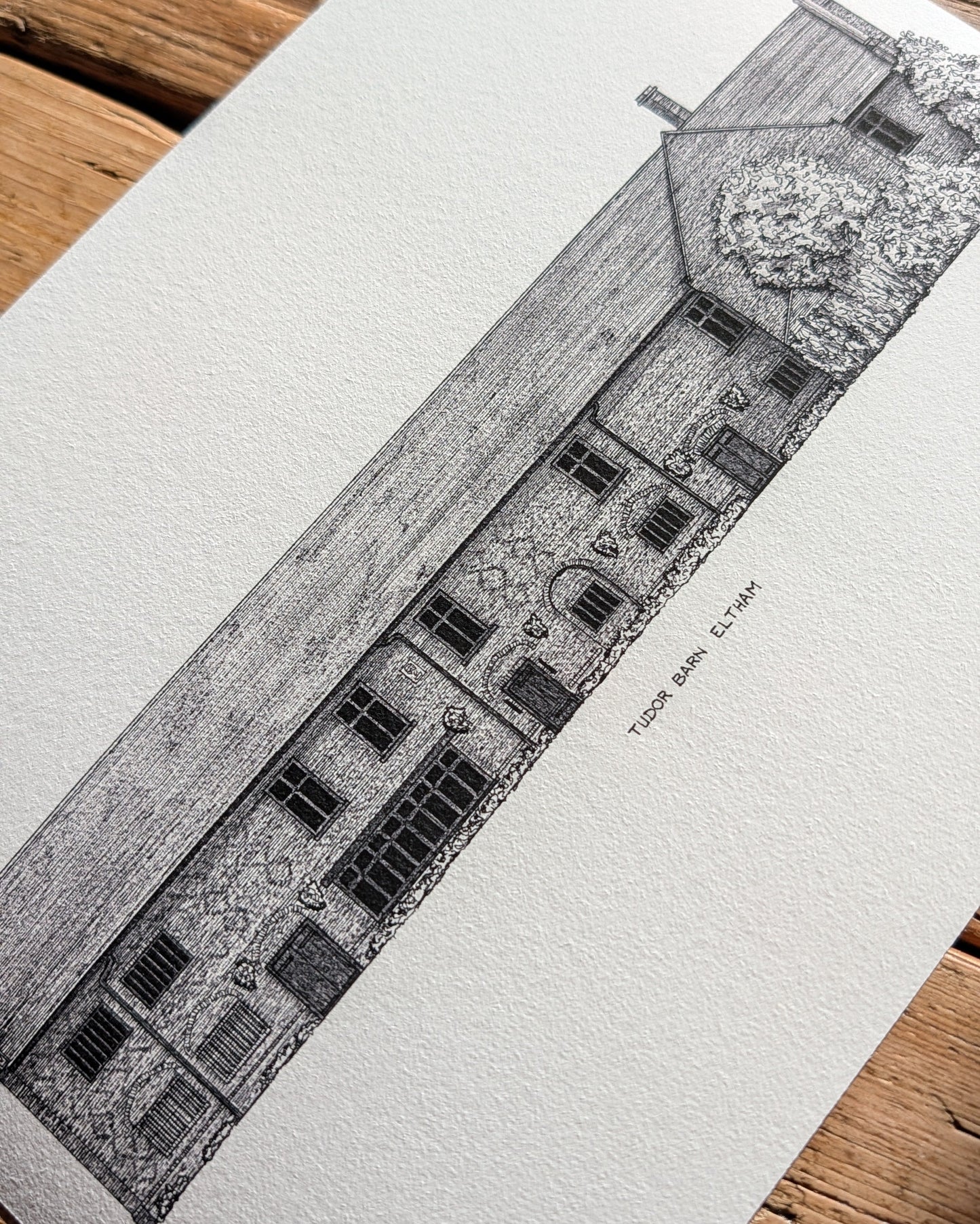 A black and white architectural sketch print of 'Tudor Barn, Eltham' on textured white paper, displayed diagonally on a rustic wooden surface.